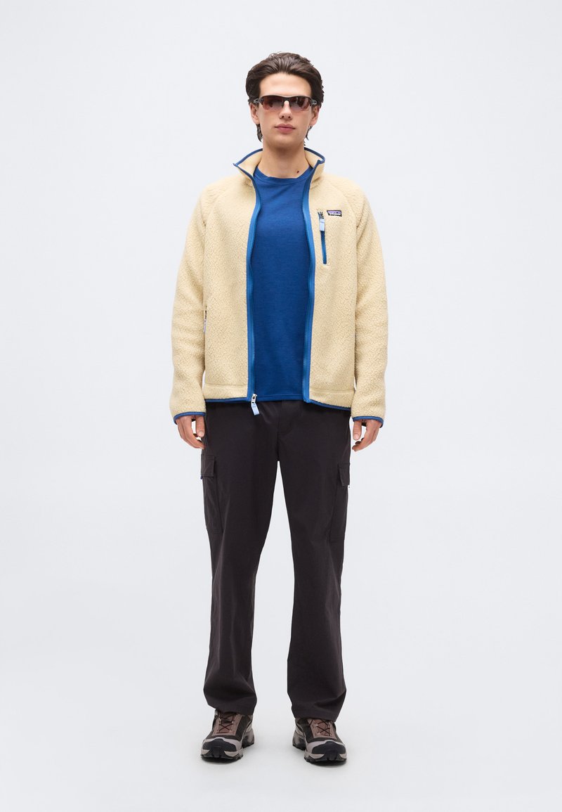 Homme portant une veste en polaire beige, une chemise bleue, un pantalon cargo noir, des chaussures de randonnée et des lunettes de soleil, debout devant un fond clair uni.