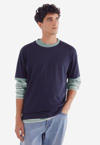 Mörkblå kortärmad skjorta lager över en ljusgrön långärmad skjorta med vita ränder, tillsammans med ljusblå jeans. Slät textur.