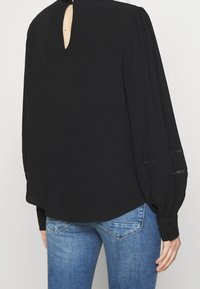 Sort bluse med lange, puffede ærmer. Har en nøglehulslukning bagpå, teksturerede detaljer ved manchetterne og en rund nederkant. Bæres med blå jeans.