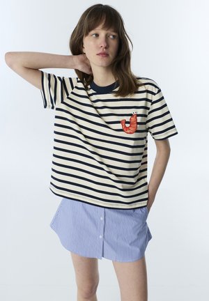 Joven mujer que lleva una camisa de rayas blancas y negras con un parche rojo de camarón, combinada con una falda azul de rayas con botones.