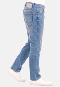 Jeans de mezclilla azul claro con corte recto, diseño de cinco bolsillos, parche con logo en la parte trasera y detalles de costura sutiles; combinados con zapatillas blancas.