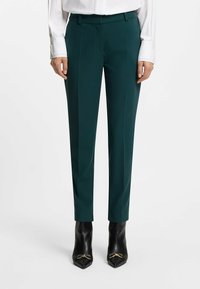 Maßgeschneiderte dunkelgrüne Hose mit glatter Textur, flachem Bund und seitlichen Taschen, kombiniert mit spitzen schwarzen High Heels.