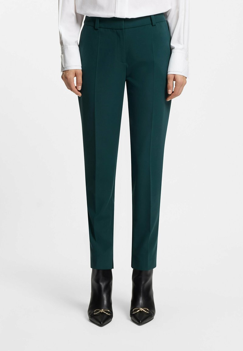Maßgeschneiderte dunkelgrüne Hose mit glatter Textur, flachem Bund und seitlichen Taschen, kombiniert mit spitzen schwarzen High Heels.