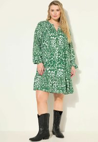 Robe verte à motifs avec des motifs blancs, manches longues, décolleté en V et ourlet évasé. Associée à des bottes de cowboy noires. Texture de tissu lisse.