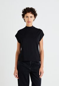 Svart kortärmad topp med hög krage, slät vaddering, avslappnad passform och diskret axeldesign, i kombination med svarta jeans.
