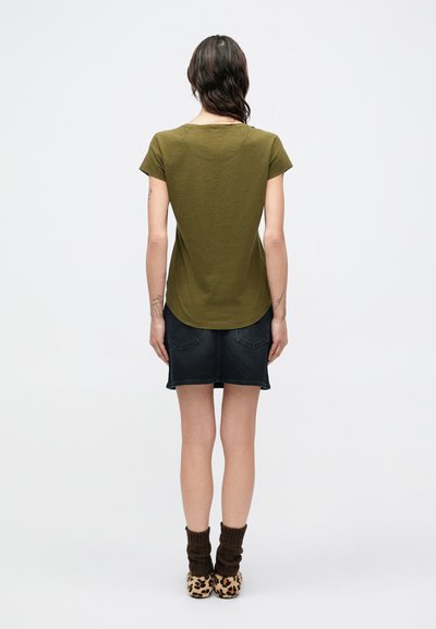 Κοντομάνικο πουκάμισο σε ελιά πράσινο, με στρογγυλεμένη γωνία στη βάση, συνδυασμένο με σκουρόχρωμη τζιν φούστα και παπούτσια με λεοπάρ εκτύπωση. Η υφή φαίνεται απαλή και casual.