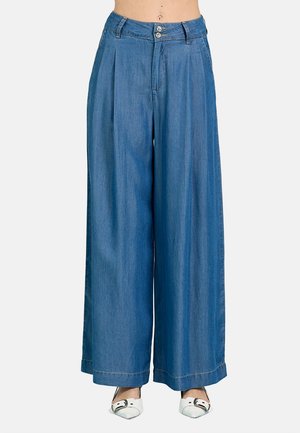 Pantaloni in denim a vita alta e gamba larga con pieghe frontali, chiusura con due bottoni, passanti per cintura e scarpe bianche appuntite.