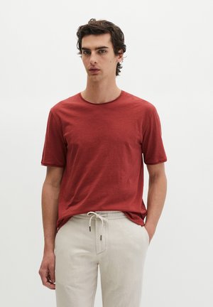 Junger Mann mit dunklen Haaren, der ein kurzärmeliges T-Shirt in Rostrot und eine hellbeige Hose mit Kordelzug vor einem einfarbigen weißen Hintergrund trägt.