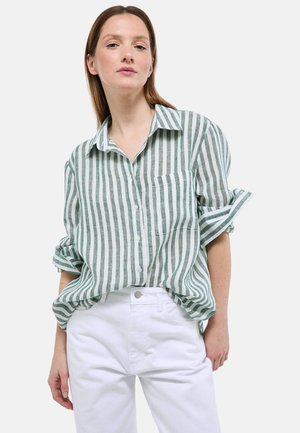 Femme portant une chemise à rayures verticales vertes et blanches avec les manches retroussées, associée à un pantalon blanc taille haute, debout avec confiance.