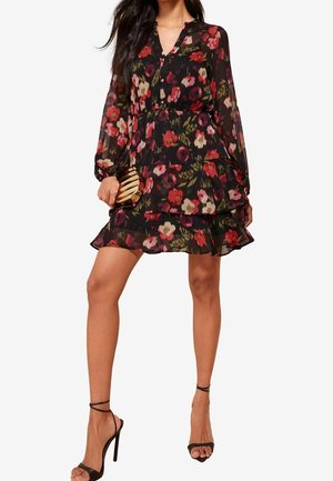 Woman wearing black floral long-sleeve tiered mini dress with ruffled hem, holding a small striped clutch, and black high-heeled sandals.