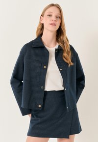 Navy-blauwe gestreepte blazer met goudkleurige hardware en opgestikte zakken, gecombineerd met een bijpassende gestreepte minirok. Witte T-shirt eronder.