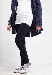 Parka azul-marinho com fecho de correr, túnica branca, jeans skinny azul-escuro e ténis cinzentos com solas brancas. Textura suave, design casual.