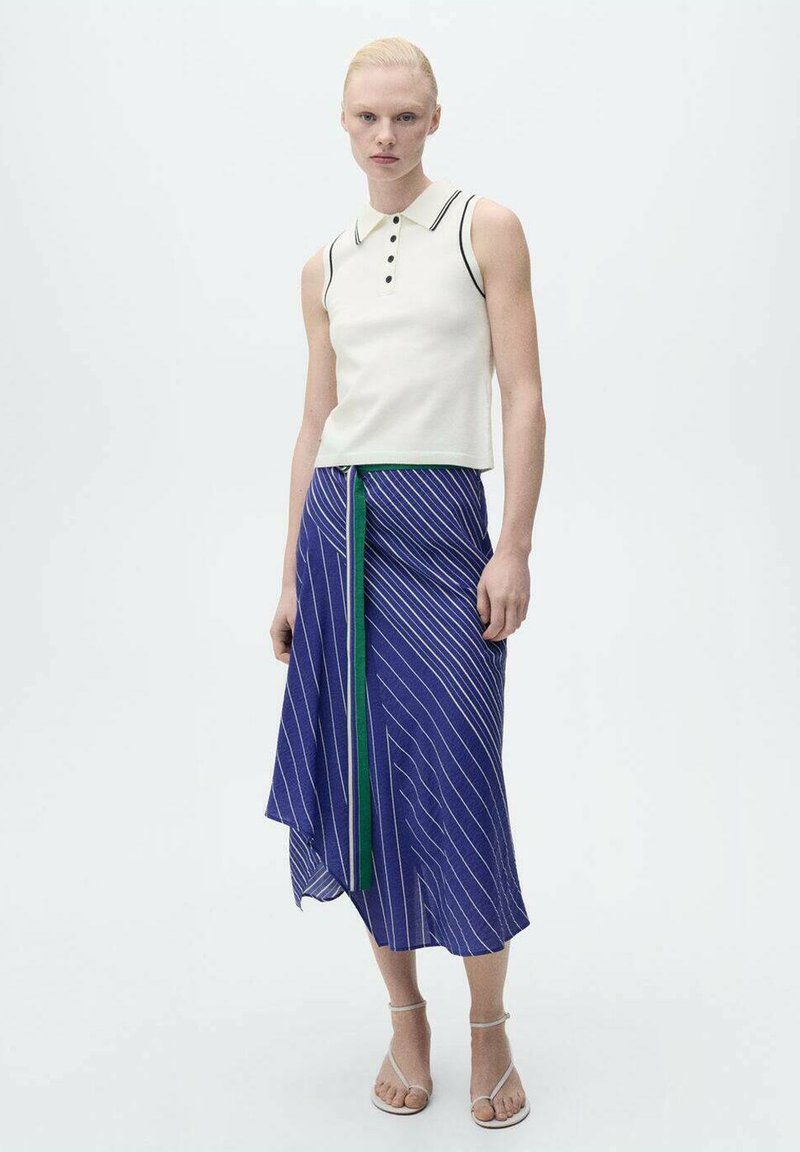 Haut polo sans manches blanc avec accents noirs, associé à une jupe rayée bleue avec des détails verts, et des sandales claires.