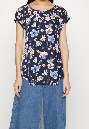 Femme portant un chemisier bleu marine à manches courtes avec un imprimé floral rose, blanc et bleu, associé à un jean en denim bleu à jambes larges.