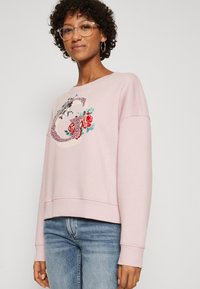 Sudadera rosa claro con diseño bordado de flores y letras, cuello redondo, mangas largas y dobladillo acanalado. Combinada con jeans azules.