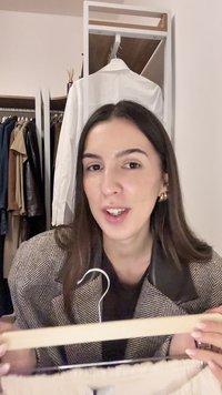 Žena s dlouhými tmavými vlasy a zlatými kruhovými náušnicemi drží dřevěný ramínko s oblečením před skříní plnou šatstva.