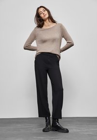 Jasnobrązowy sweter z długim rękawem o luźnym kroju, w połączeniu z czarnymi szerokimi spodniami i czarnymi masywnymi butami, na neutralnym tle.