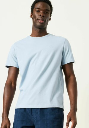 Muž ve světlomodrém tričku s krátkým rukávem a tmavě modrých texturovaných kalhotách stojící proti jednobarevnému pozadí.