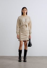 Φόρεμα φούτερ σε μπεζ χρώμα με λεπτομέρειες ραφές, λογότυπο Calvin Klein στο στήθος. Το μοντέλο φοράει μαύρες μπότες μέχρι το γόνατο και κρατάει μια μικρή μαύρη τσάντα.