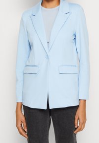Blazer bleu clair en tissu doux, avec une fermeture à un bouton, des revers à encoche et deux poches avant. Coupe droite.