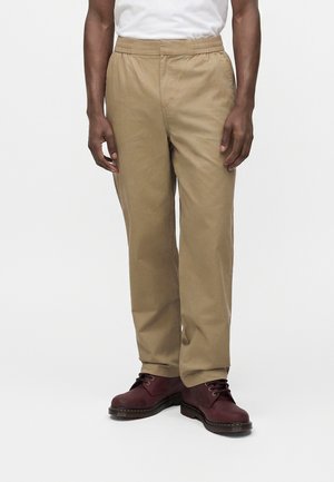 Personne portant un pantalon beige droit avec taille élastique et des bottes à lacets rouge foncé, debout sur un fond blanc.