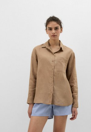 Jonge vrouw met donker haar in een losse knot draagt een lichtbruine blouse met knopen en lange mouwen en blauw-wit gestreepte korte broek tegen een effen achtergrond.