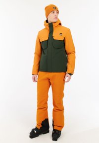 Oranžová a zelená nepremokavá lyžiarska bunda s dvoma veľkými prednými vreckami; v kombinácii s oranžovými lyžiarskymi nohavicami a čiernymi lyžiarskymi topánkami. Doplnková pletená oranžová čiapka.