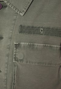 Zbliżenie na kieszeń oliwkowej kurtki z klapą, widocznym ściegiem dekoracyjnym i ciemnym guzikiem blisko kołnierza.