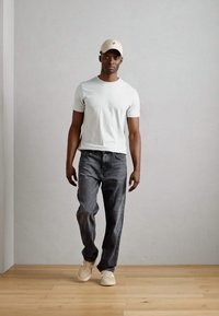 Camiseta gris claro, jeans gris oscuro y zapatillas beige. Una gorra beige completa el conjunto. El fondo presenta una pared y un suelo simples y neutros.