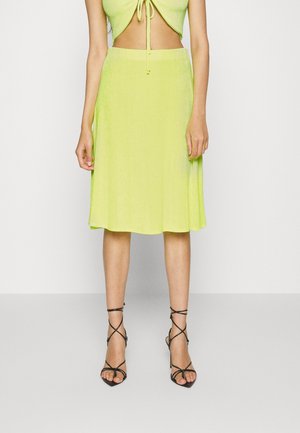 Kvinde iført limegrøn knælængde nederdel og matchende top med sorte, remsatte højhælede sandaler, stående mod en lys baggrund.