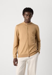 Mannelijke model met een donkere huid en kort haar, gekleed in een beige ritsvest en witte broek, staand tegen een effen lichte achtergrond.