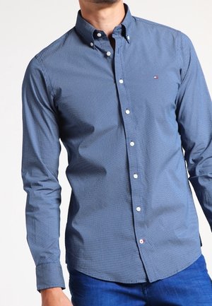 Uomo che indossa una camicia azzurra aderente a maniche lunghe con un piccolo logo sul petto e jeans blu.