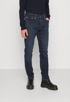 Jeans em denim azul escuro, ajustados, com um design de cinco bolsos, apresentando rebites em tom cobre, costuras em contraste e uma textura de tecido suave.