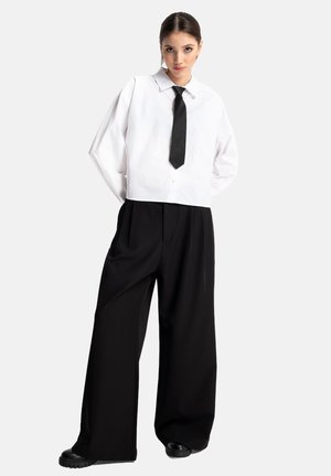 Skrojona biała koszula z kołnierzykiem i guzikami, w połączeniu z luźnymi czarnymi spodniami. Uzupełniona czarnym krawatem i teksturowanym czarnym obuwiem.