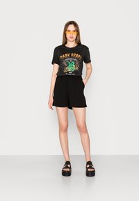T-shirt noir avec un imprimé graphique coloré, associé à un short noir. Comprend des lunettes de soleil jaunes et des sandales noires épaisses. Fond minimaliste.