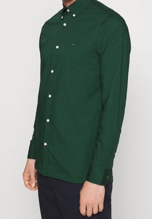 Man wearing dark green button-up shirt with small logo on chest and dark pants, standing against plain light background.