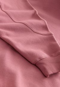 Sweat-shirt rose en tissu texturé, avec des poignets côtelés et une surface lisse, présentant des détails de coutures subtils pour un intérêt de design supplémentaire.
