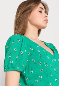 Vestido verde com mangas bufantes, apresentando um padrão floral em branco e rosa. Tem um decote em V e tecido texturizado.