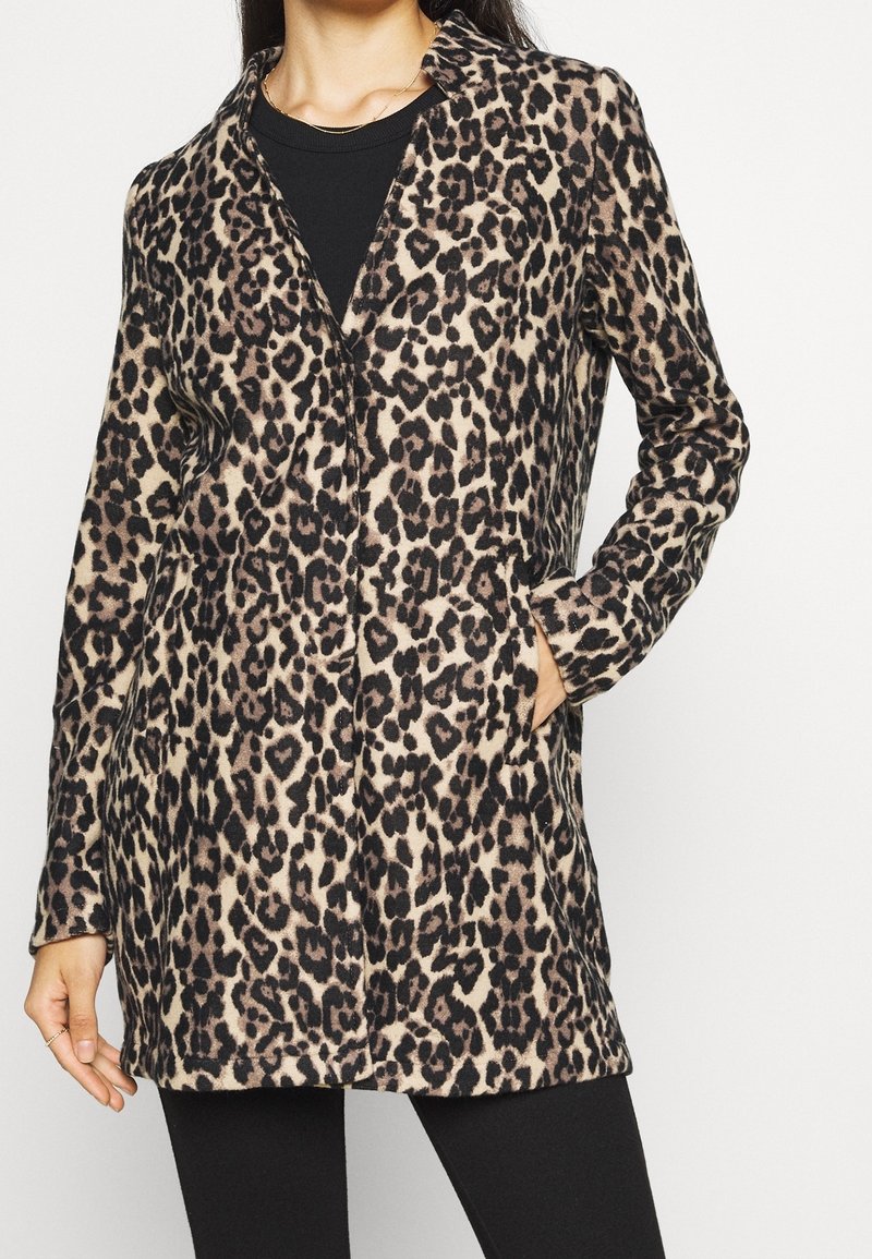 Kobieta w długim rękawie, w leopardzim płaszczu, z czarną bluzką i czarnymi spodniami, z jedną ręką w kieszeni płaszcza.