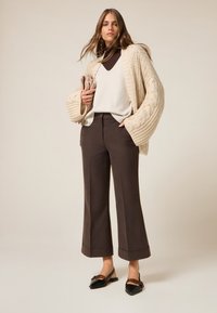 Cardigan de punto beige, suéter crema y pantalones de pierna ancha marrón oscuro, combinados con planos negros de punta afilada con un acento de cadena.