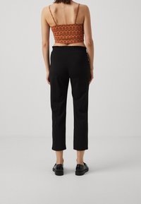 Femme portant un pantalon court noir, des chaussures noires et un crop top orange à motifs sans manches, debout de dos sur un fond uni.