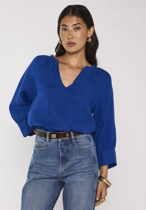 Donna che indossa una camicetta blu a maniche lunghe con scollo a V, infilata in jeans blu a vita alta con cintura marrone, indossa gioielli in oro.