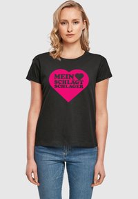 Sort bomulds t-shirt med rund halsudskæring, korte ærmer og et pink hjerte design med fed sort tekst, der står "MEIN ♥ SCHLÄGT SCHLAGER."