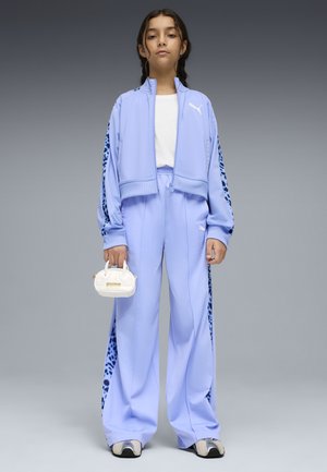 Ung person iført lyse lilla Puma træningsdragt med leopardprintstriber, hvid t-shirt, sneakers og lille hvid Puma håndtaske, stående og vendt fremad.