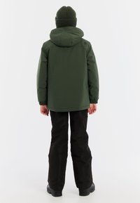 Grüne wasserfeste Jacke mit Kapuze, elastischen Bündchen, kombiniert mit schwarzen Hosen und Stiefeln. gestrickte grüne Mütze auf dem Kopf.