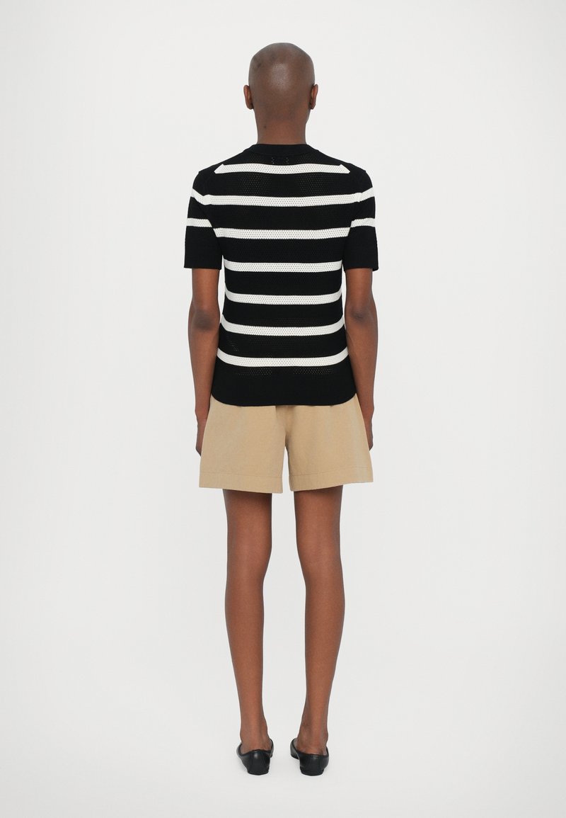 Pull en tricot à rayures noires et blanches avec des manches courtes, associé à un short beige et des chaussures noires à enfiler, vu de dos.