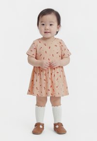 Copil mic în picioare, purtând o rochie portocalie cu flori, șosete albe până la genunchi și pantofi maro, zâmbind cu mâinile împreunate.