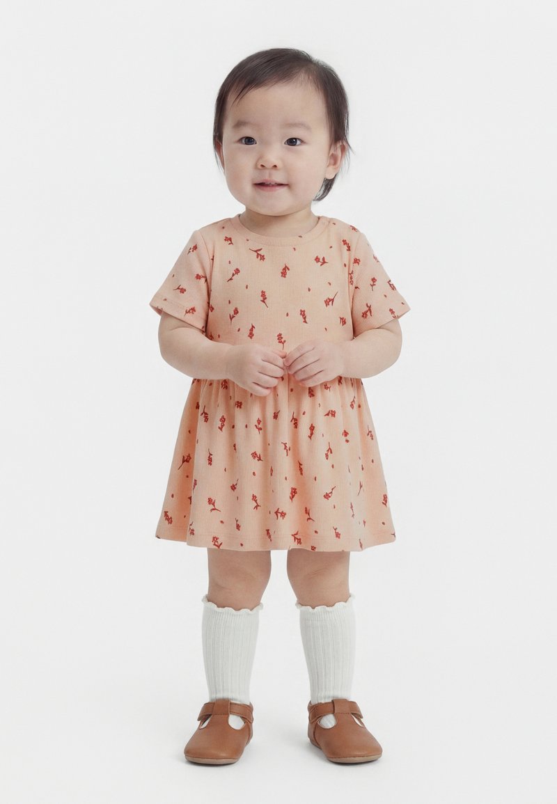 Copil mic în picioare, purtând o rochie portocalie cu flori, șosete albe până la genunchi și pantofi maro, zâmbind cu mâinile împreunate.