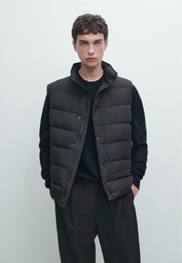 Giovane uomo con gilet trapuntato nero sopra una maglietta nera a maniche lunghe e pantaloni neri, in piedi contro uno sfondo grigio chiaro uniforme.