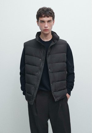 Giovane uomo con gilet trapuntato nero sopra una maglietta nera a maniche lunghe e pantaloni neri, in piedi contro uno sfondo grigio chiaro uniforme.